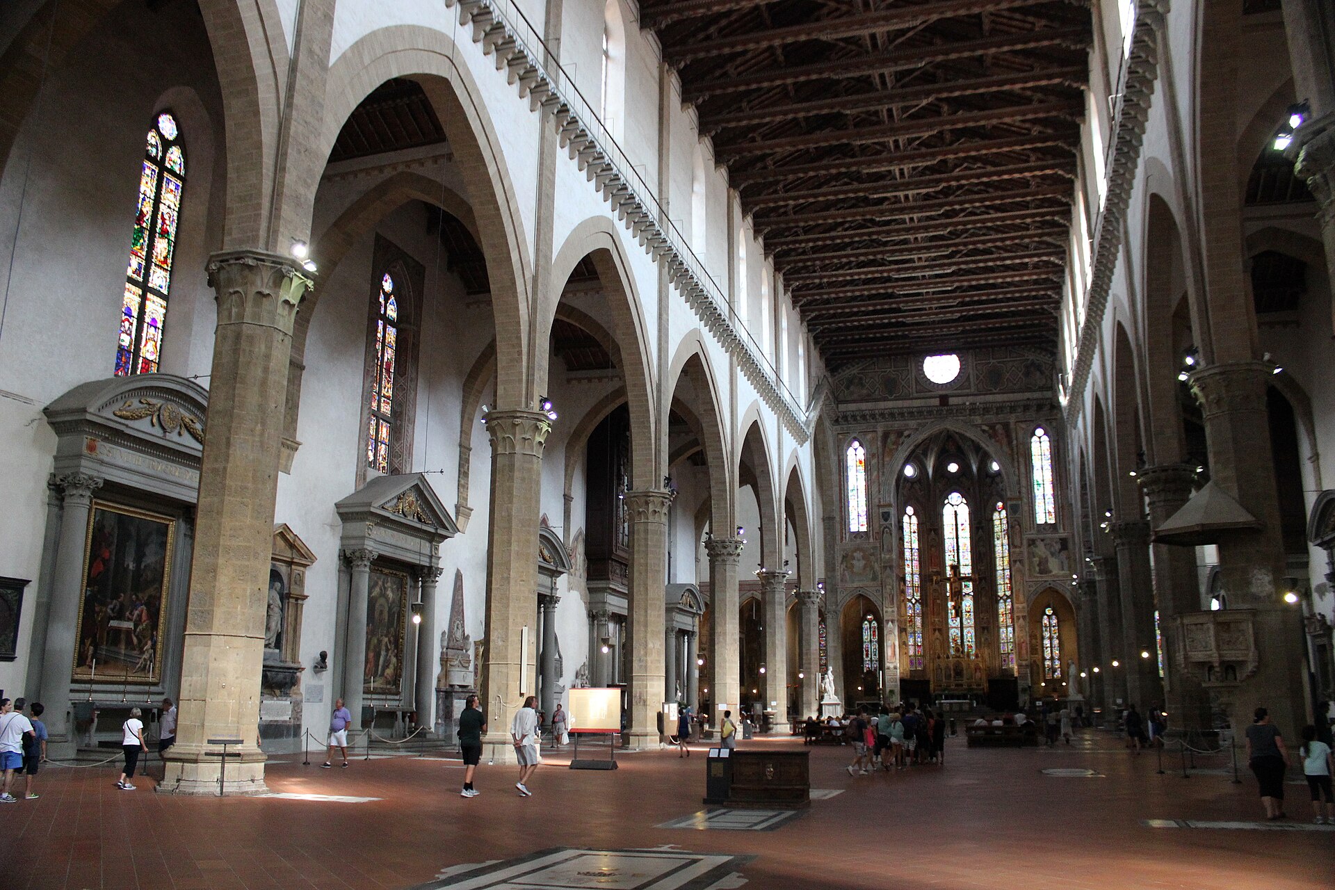 Visitors walk beneath pointed arches and a timber roof beside wall tomb monuments in the nave of Santa Croce, Florence.