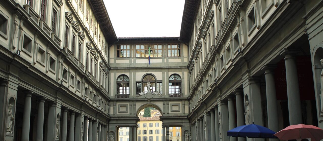 Visitors walk between the Uffizi's columned wings in Florence, with artists' displays beneath umbrellas beside the courtyard.