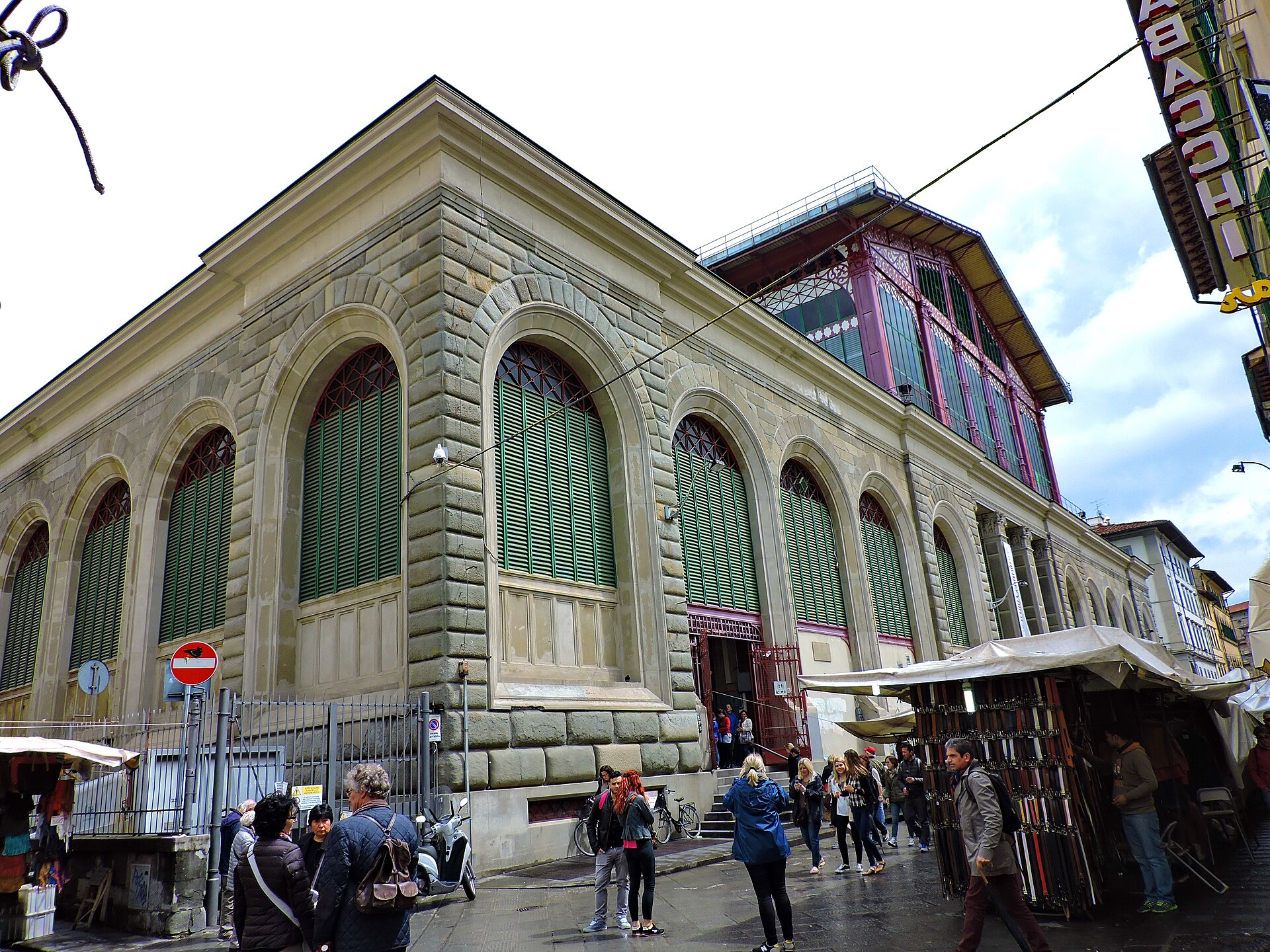 Visitors walk beside Mercato Centrale's arched windows, green shutters and red iron upper structure on a Florence market street.