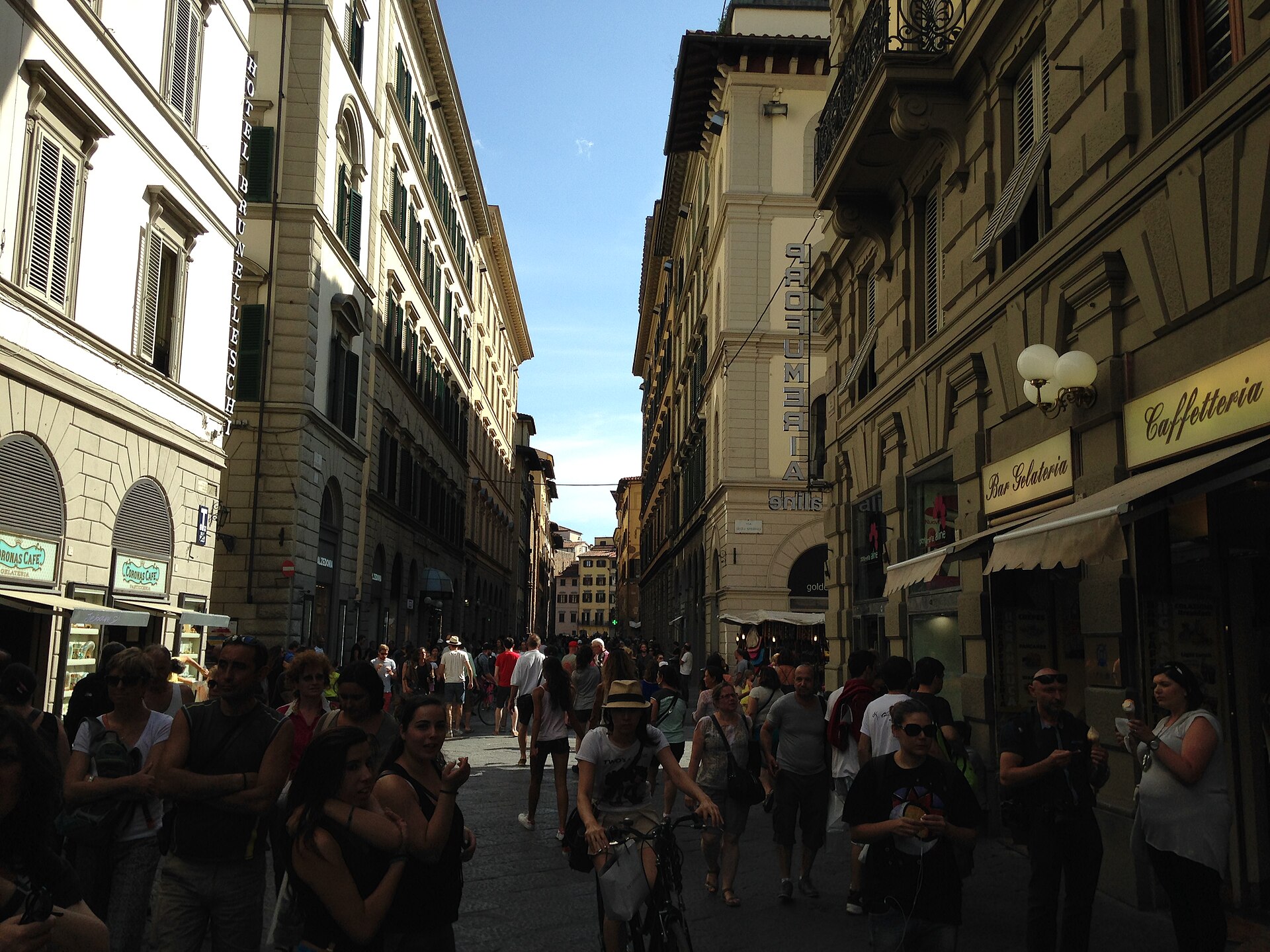 Walkers and a cyclist share Via dei Calzaiuoli in Florence beneath tall buildings with shuttered upper windows.