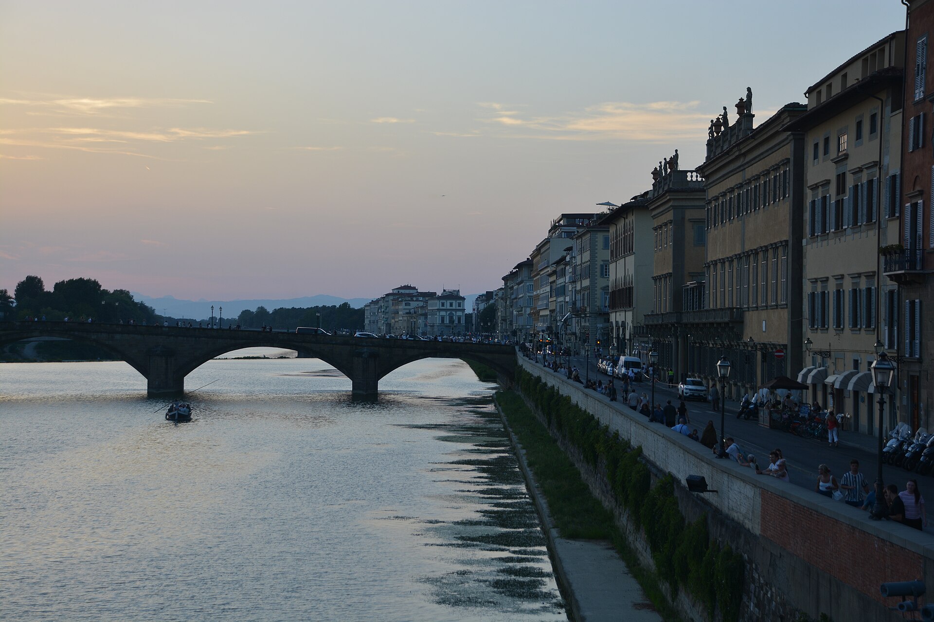 Walkers follow Florence’s Lungarno degli Acciaiuoli beside the Arno, while others sit on the parapet near Ponte Santa Trinita.