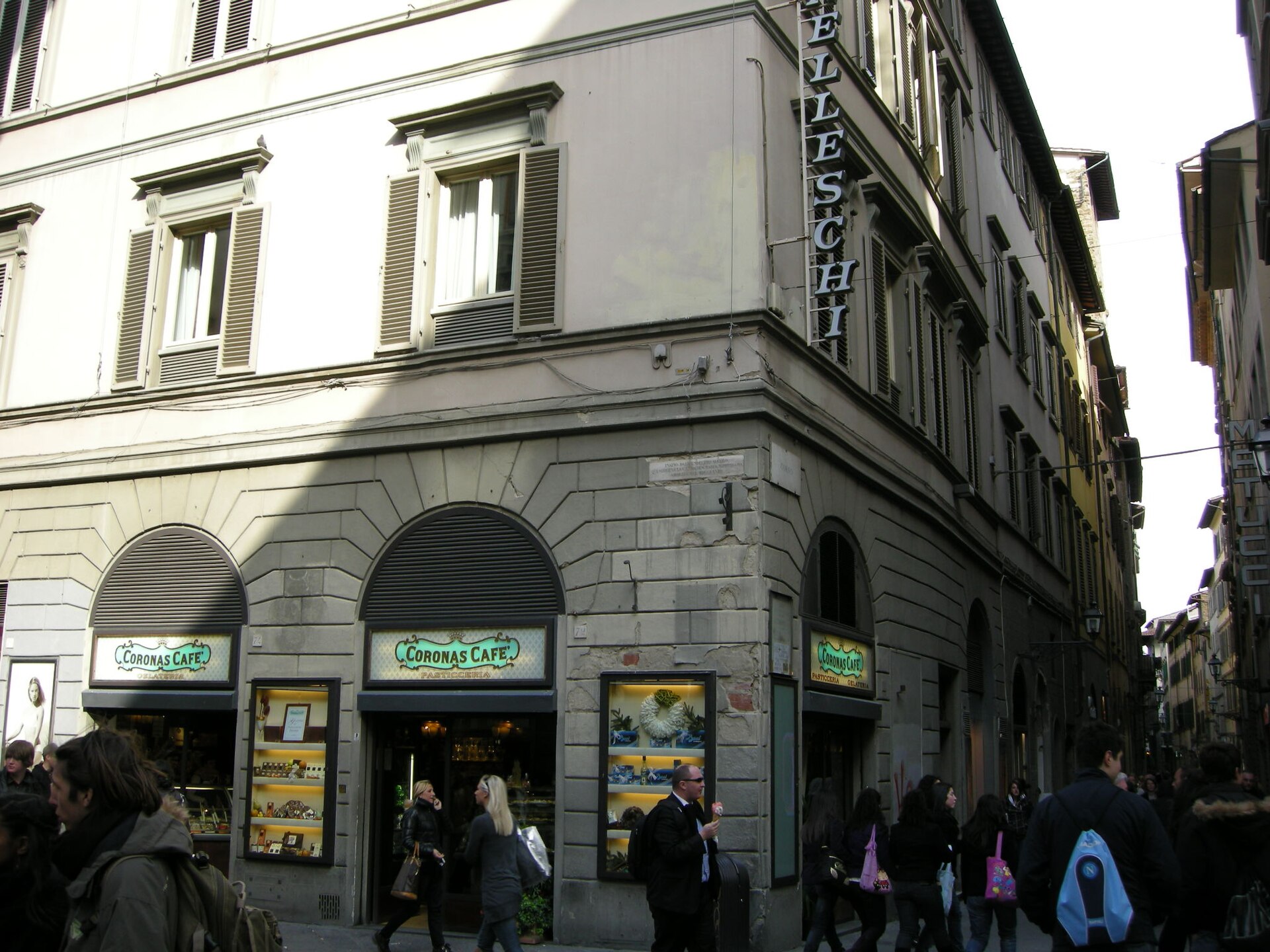 Walkers pass a stone-faced corner on Via dei Calzaiuoli in Florence, beside a narrower lane between tall buildings.