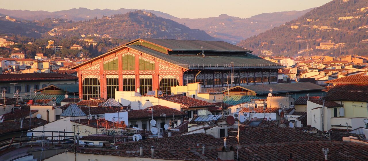 Warm evening light illuminates Mercato Centrale's iron-and-glass upper structure above Florence rooftops, with hills in the background.