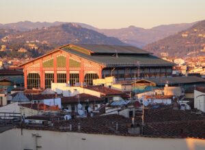 Warm evening light illuminates Mercato Centrale's iron-and-glass upper structure above Florence rooftops, with hills in the background.