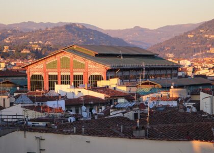 Warm evening light illuminates Mercato Centrale's iron-and-glass upper structure above Florence rooftops, with hills in the background.