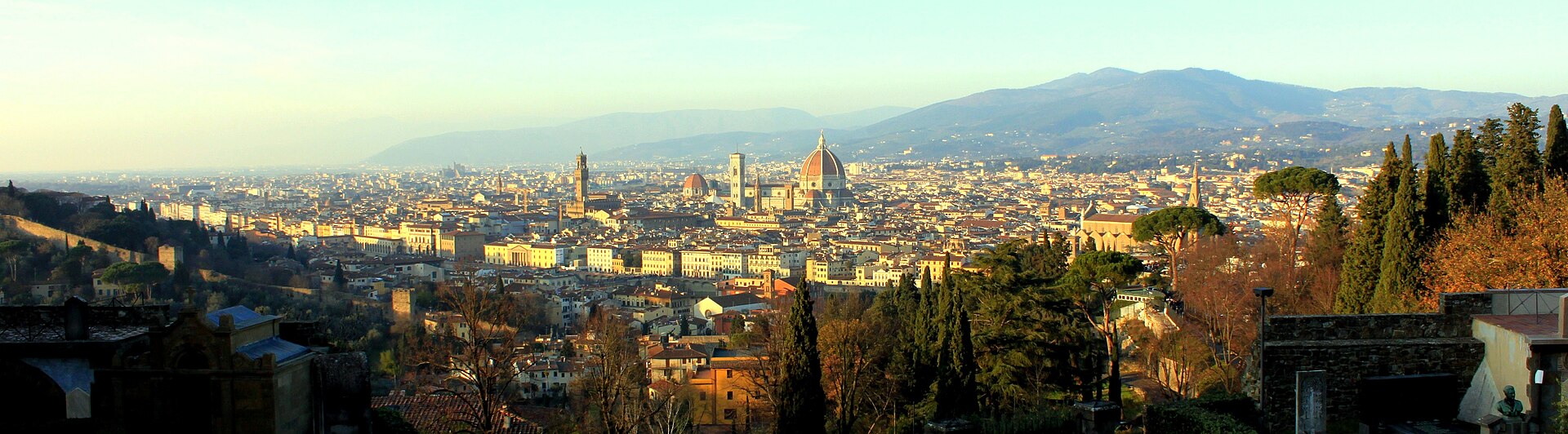 Warm light falls across Florence rooftops, with the cathedral dome and bell tower above trees and buildings below San Miniato al Monte.