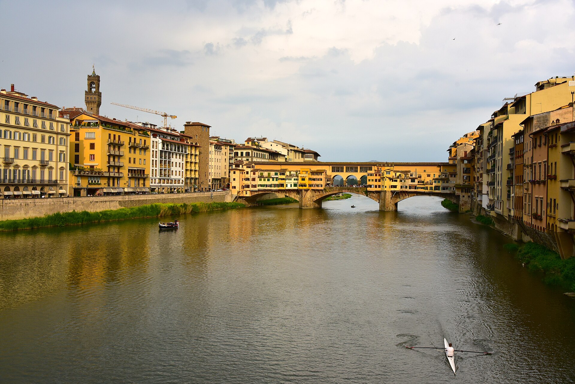Warm light falls on Ponte Vecchio and Florence’s yellow riverfront buildings as a rower and a small passenger boat cross the Arno.