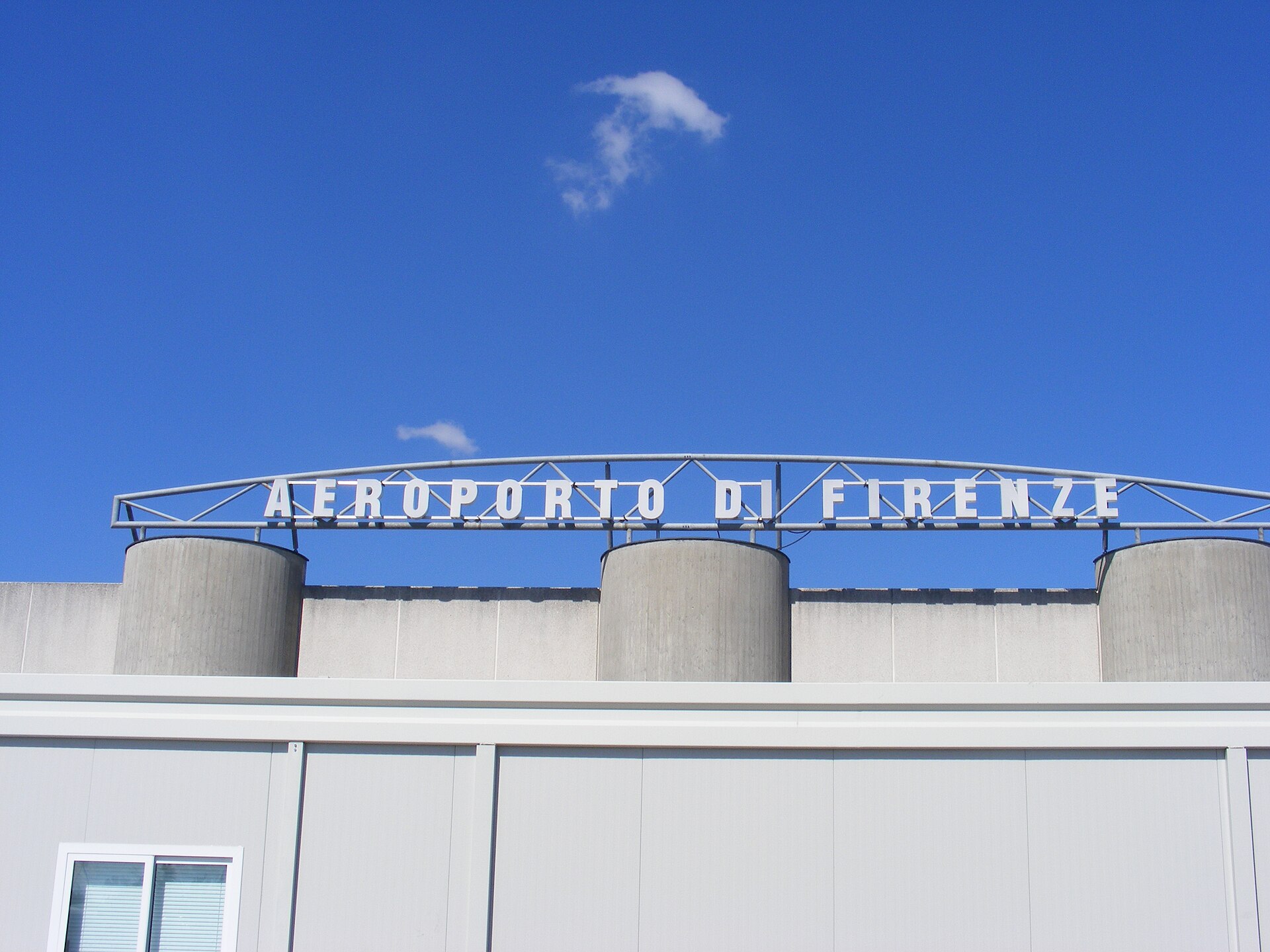 White Aeroporto di Firenze lettering stretches above the Florence Airport terminal roof beneath a blue sky.