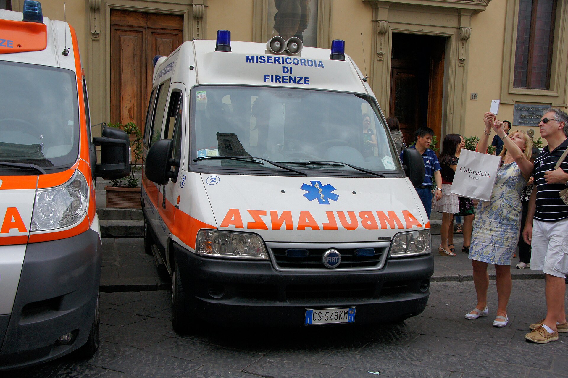 A white and orange Misericordia di Firenze ambulance with blue lights is parked beside pedestrians in Florence.