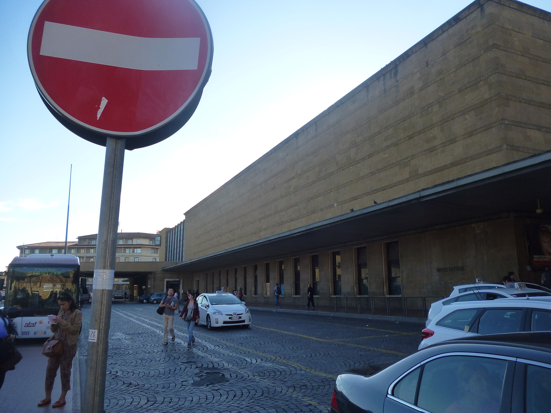 White taxis wait along the curb outside Florence Santa Maria Novella station, with pedestrians crossing the paved forecourt.