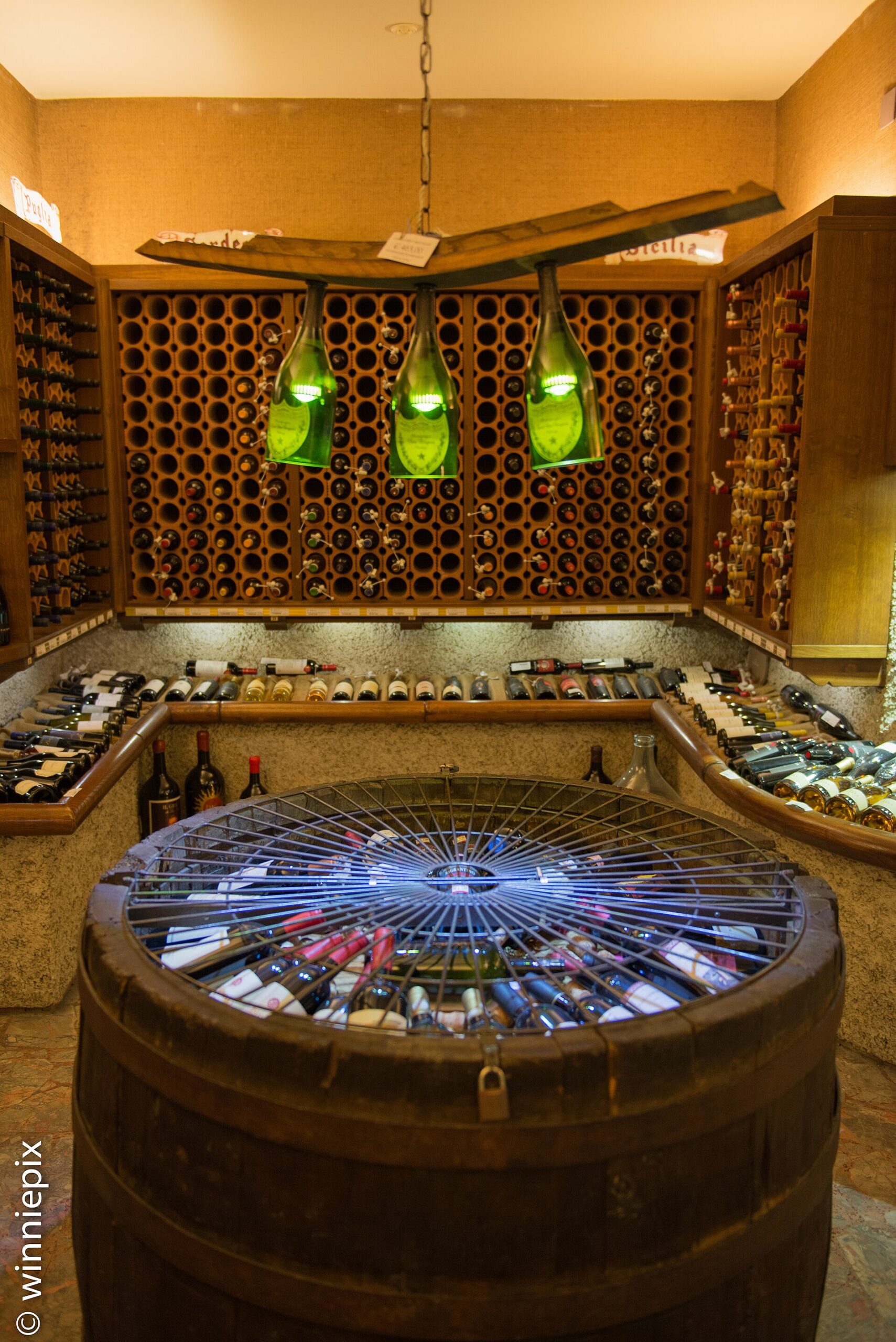 Wine bottles fill wall racks and a round barrel display beneath green bottle-shaped lamps in Florence's Enoteca Alessi cellar.