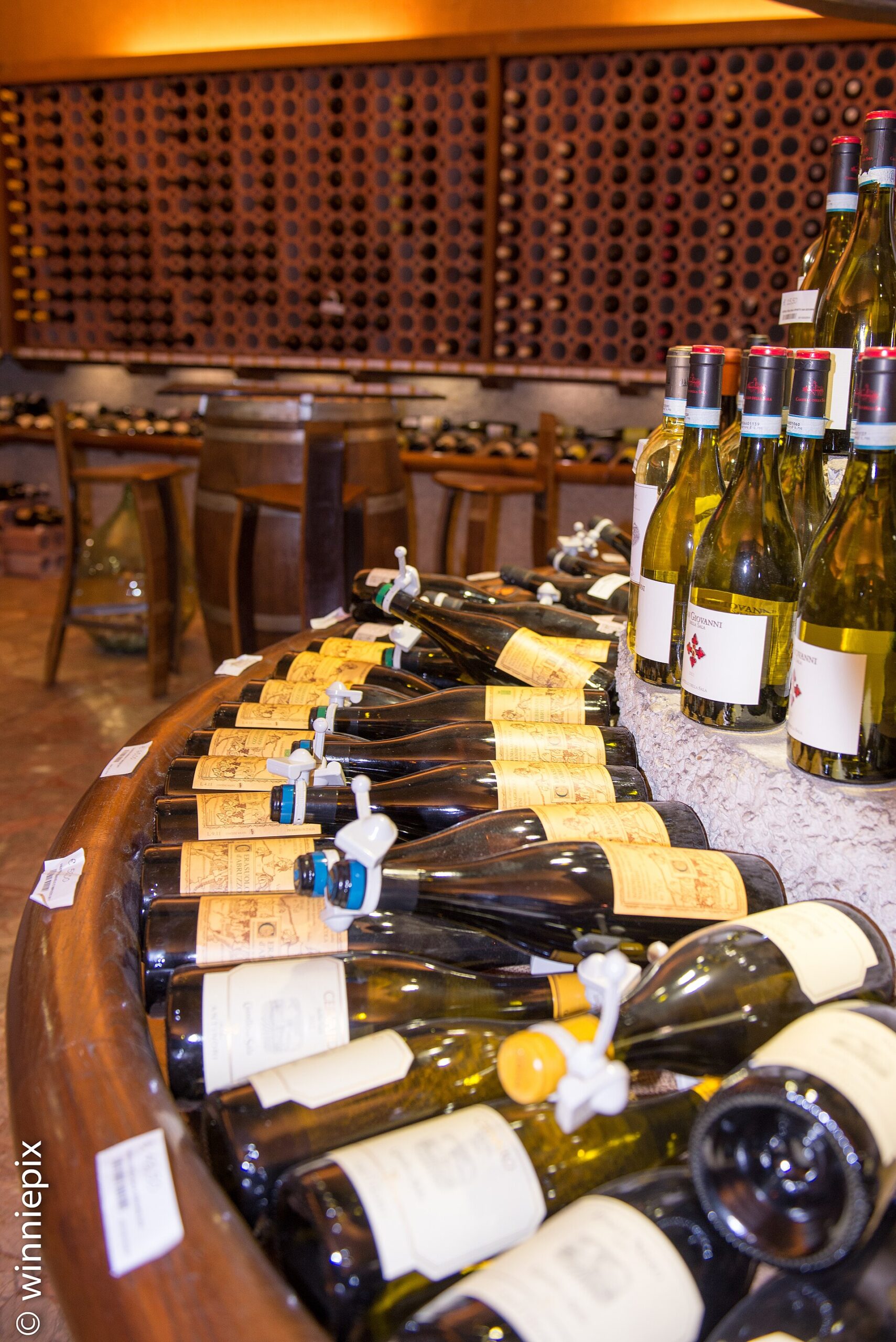 Wine bottles line a curved counter beneath bottle racks in Enoteca Alessi's Florence cellar, with high tables and stools behind.