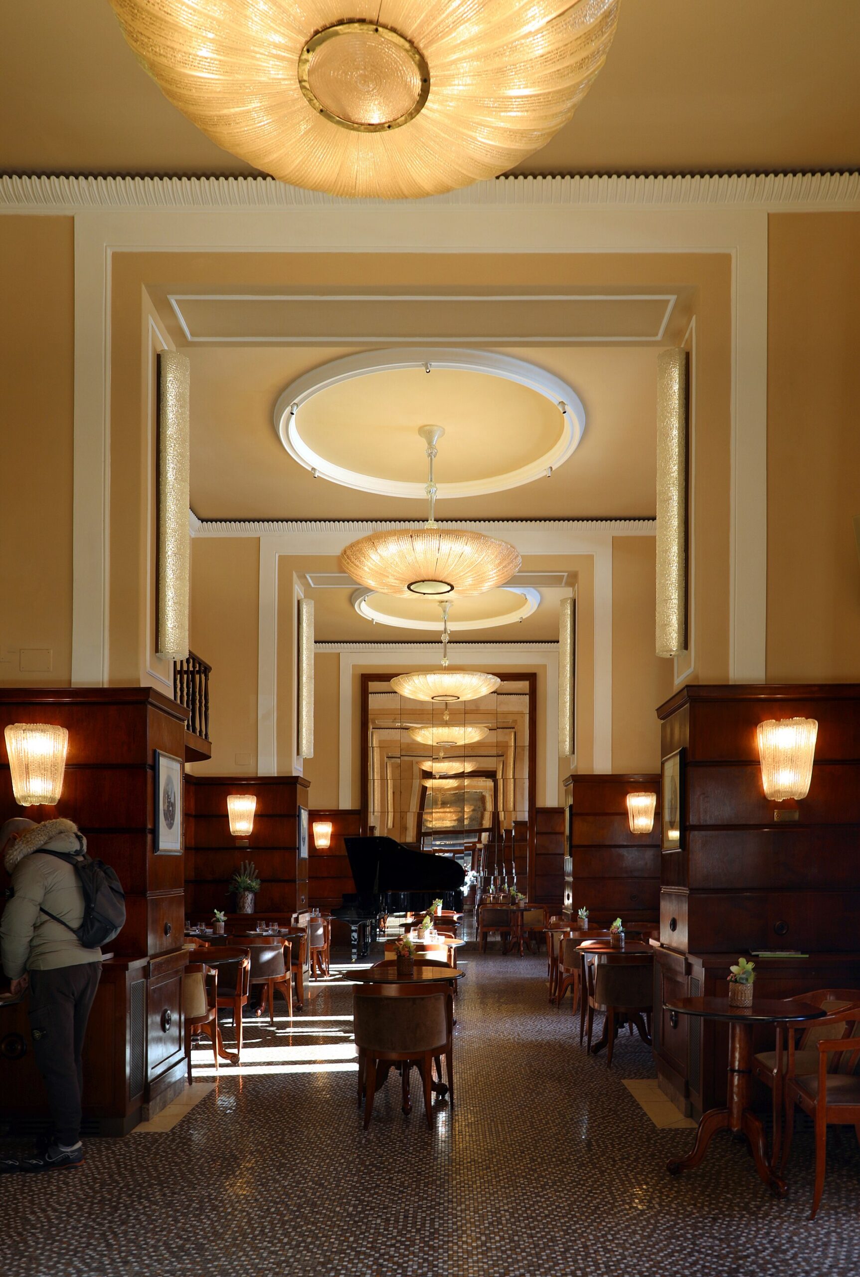 Wooden tables and chairs line Caffe Paszkowski's long Florence interior beneath large ceiling lights, with a grand piano further inside.