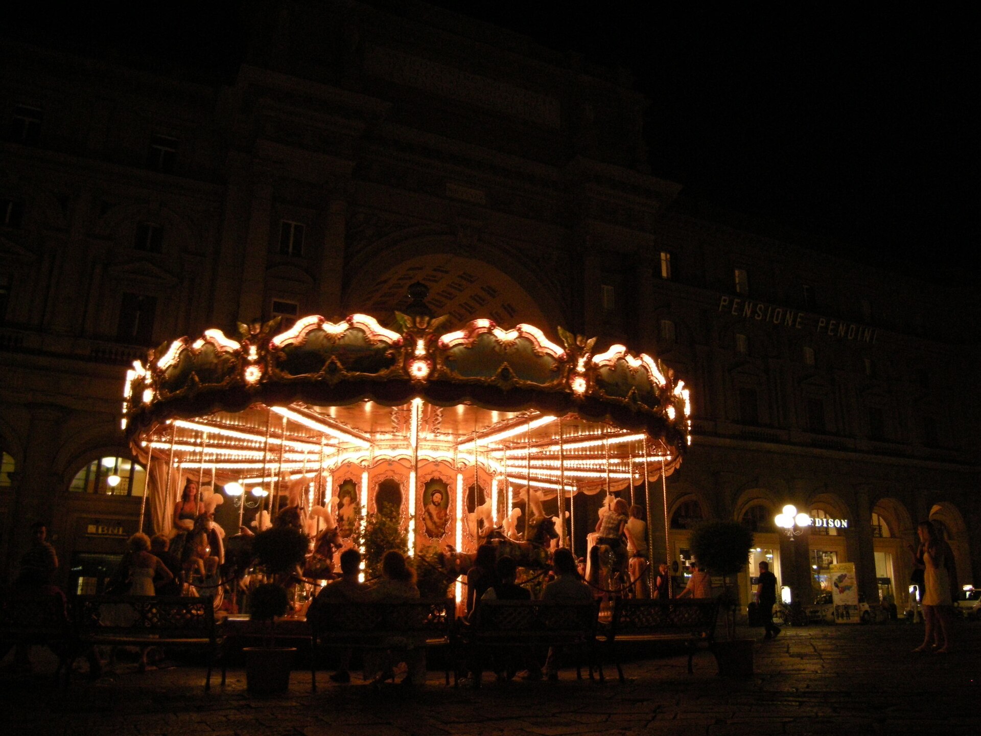 Young riders and accompanying adults gather around the illuminated carousel in Florence’s Piazza della Repubblica after dark.